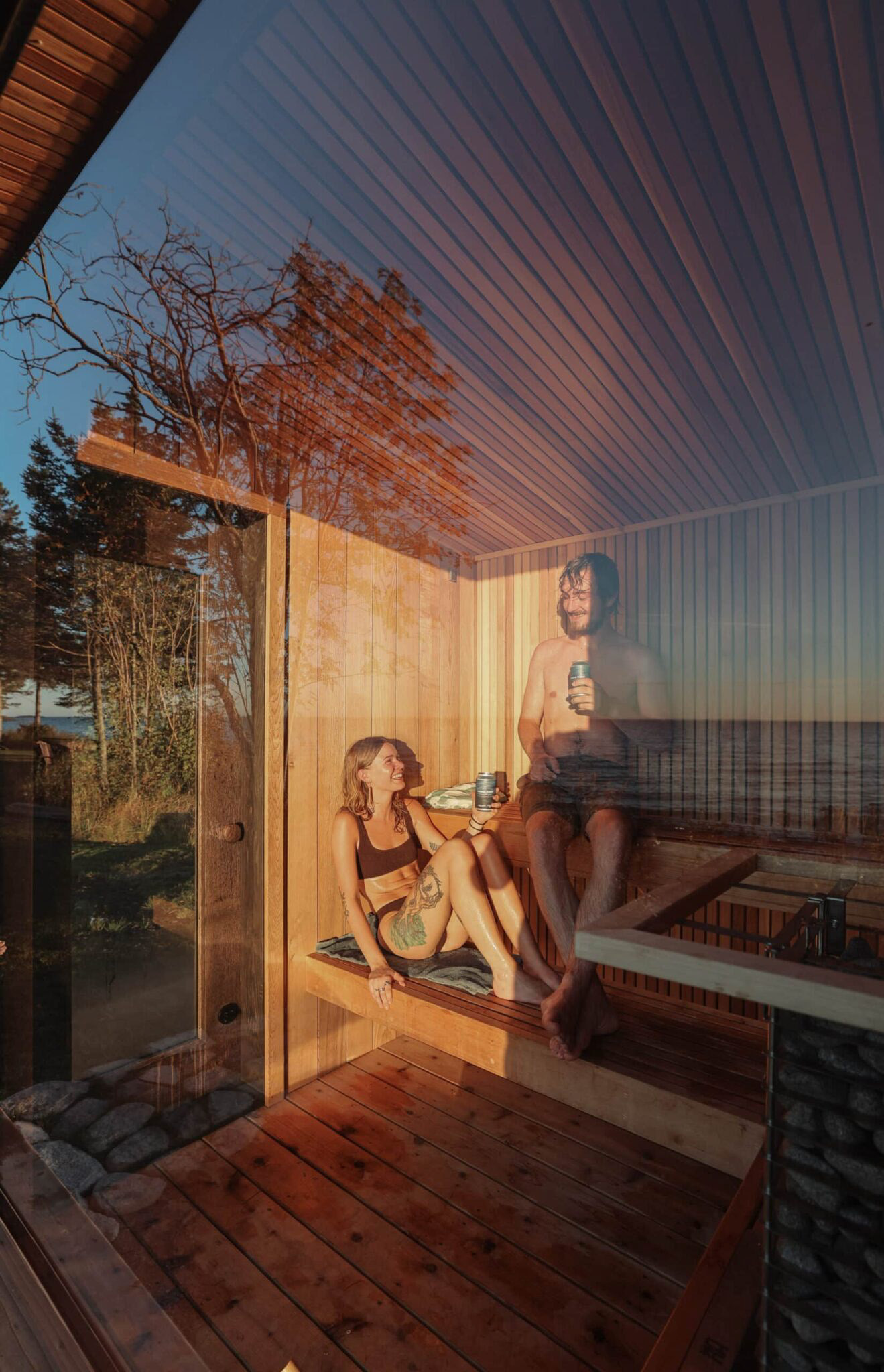 Two people relaxing on timber sauna benches in warm sunlight, seen through a window reflecting trees