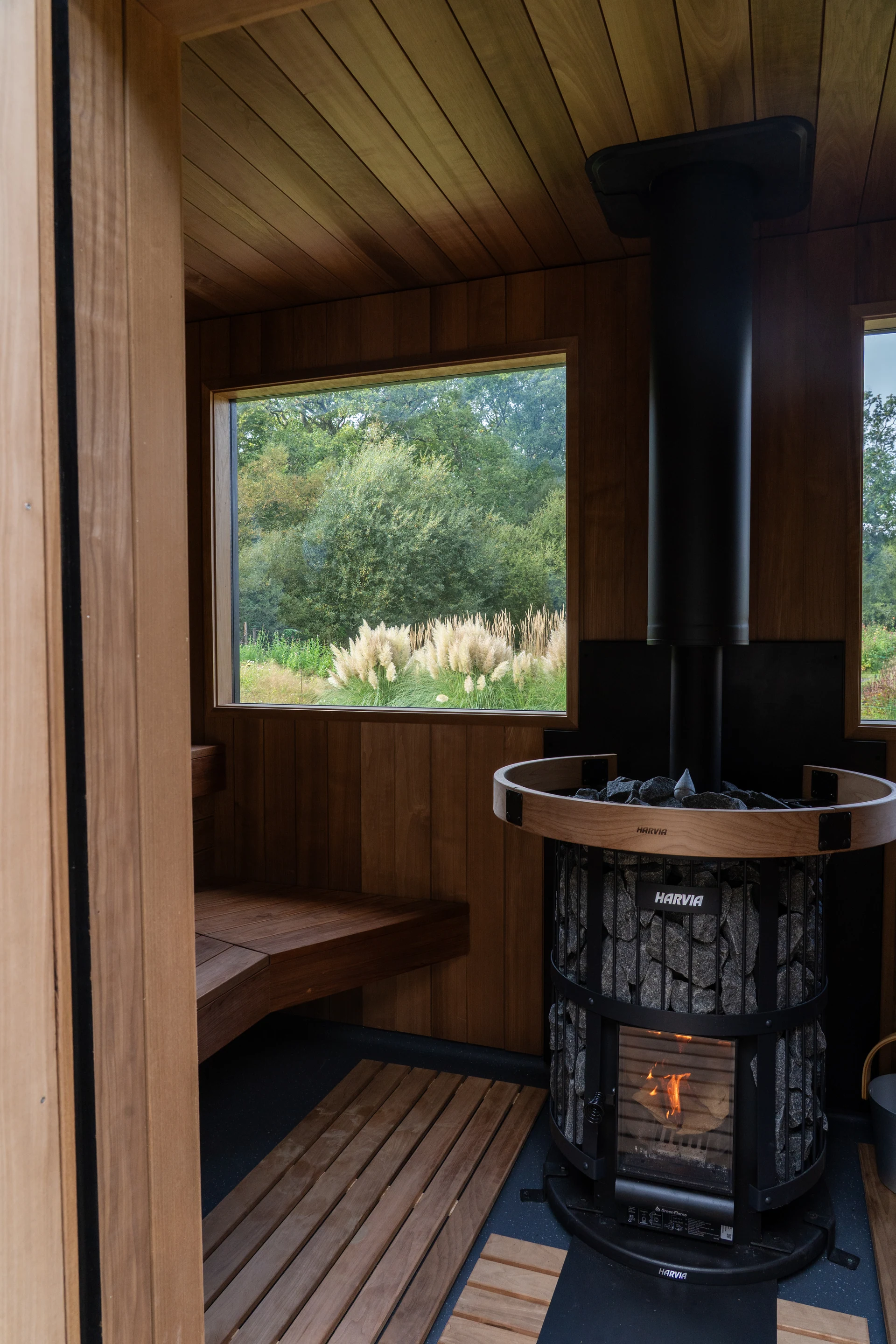 A view through the sauna doorway, with timber benches, a garden-view window and the wood-fired stove