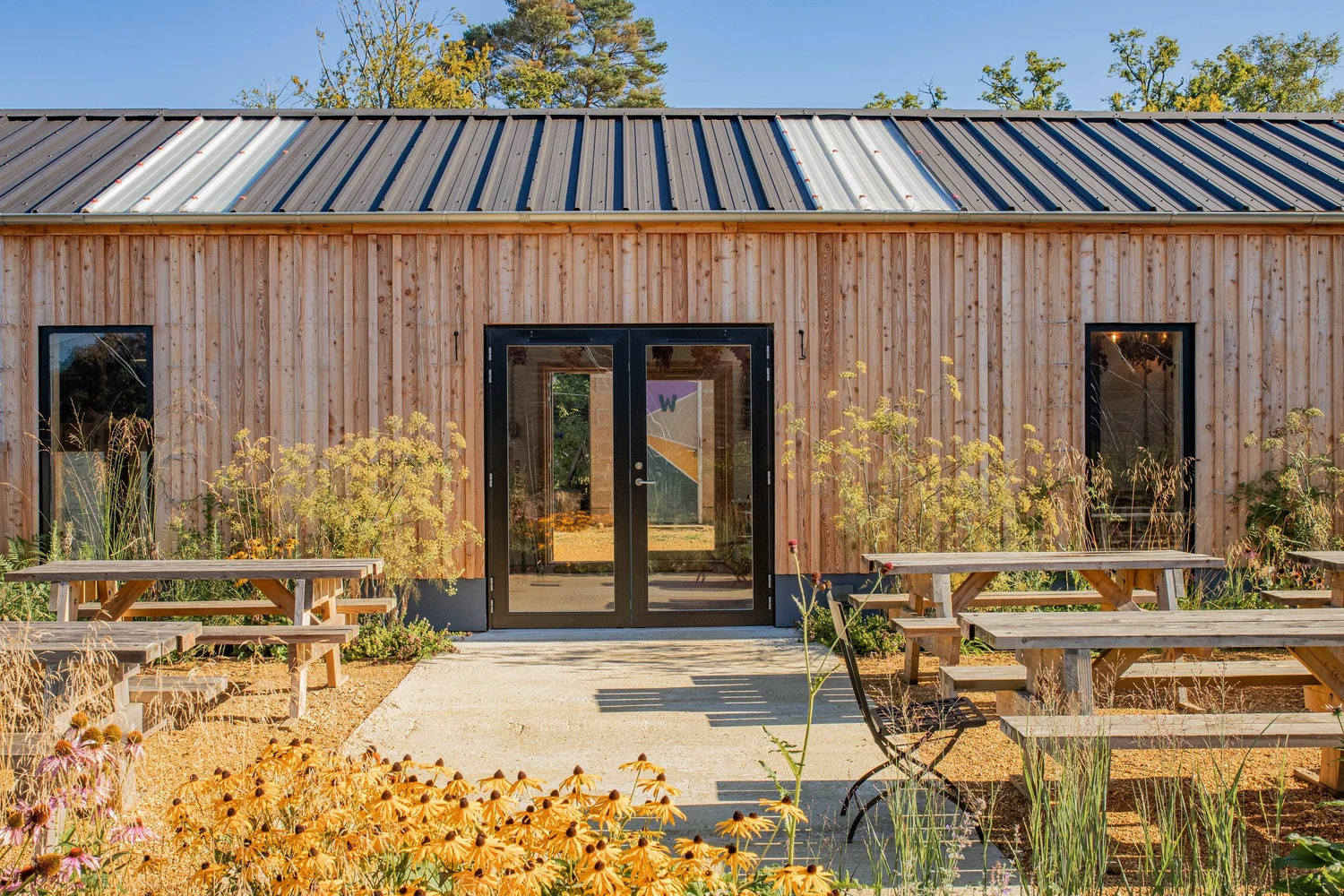 A timber-clad building with picnic benches, flowering plants and a path to the doors